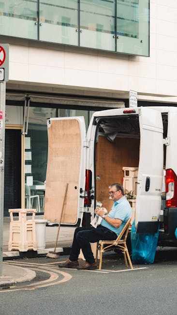 A man wearing a black cap and white t-shirt is inside a moving van, carefully handling a cardboard box during a home relocation. The van's interior shows a partially open side door, revealing outdoor