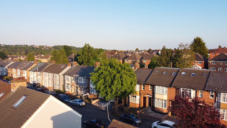 Aerial view of a residential street in Sutton during daylight, showing a row of terraced houses with various brick and painted facades, some with bay windows and dormer windows on the roof. Several parked cars line the road, including sedans and hatchbacks, with a large leafy tree situated centrally among the houses. The street appears calm and tidy, with clear skies overhead. In the foreground, a partially visible white building with a sloped roof casts a shadow on the street below. This scene depicts a typical suburban neighbourhood suitable for home relocation services, with Sutton Removals positioned to handle furniture transport, packing, and loading processes during house moves in the SM1 area.